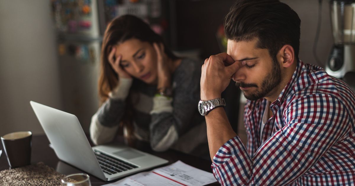 A man and woman sitting side by side, looking at a laptop and pile of bills on a table. They look stressed.